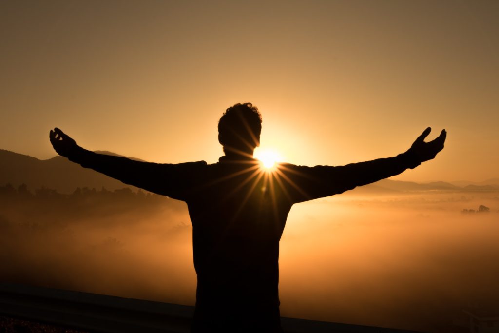 Silhouetted man spreads his arms in front of a rising sun as if to embrace it