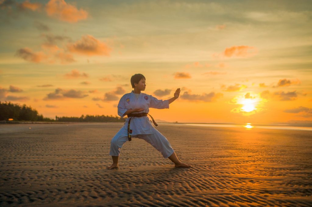 Youth in martial arts uniform stands on beach in front of sunset in martial arts stance