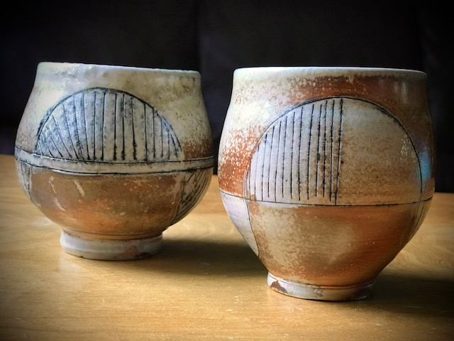Two tan and cream-colored wood-fired ceramic cups with geometric etchings sit atop a wooden surface.