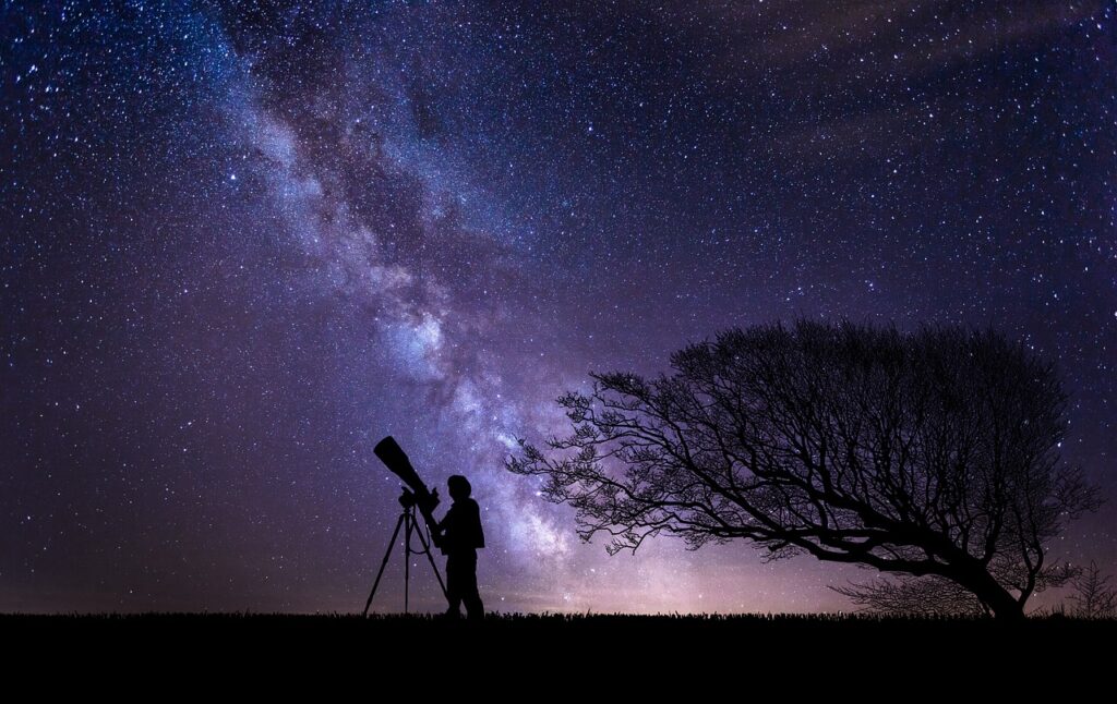 Person with large telescope stands silhouetted in front of night sky with milky way in the background