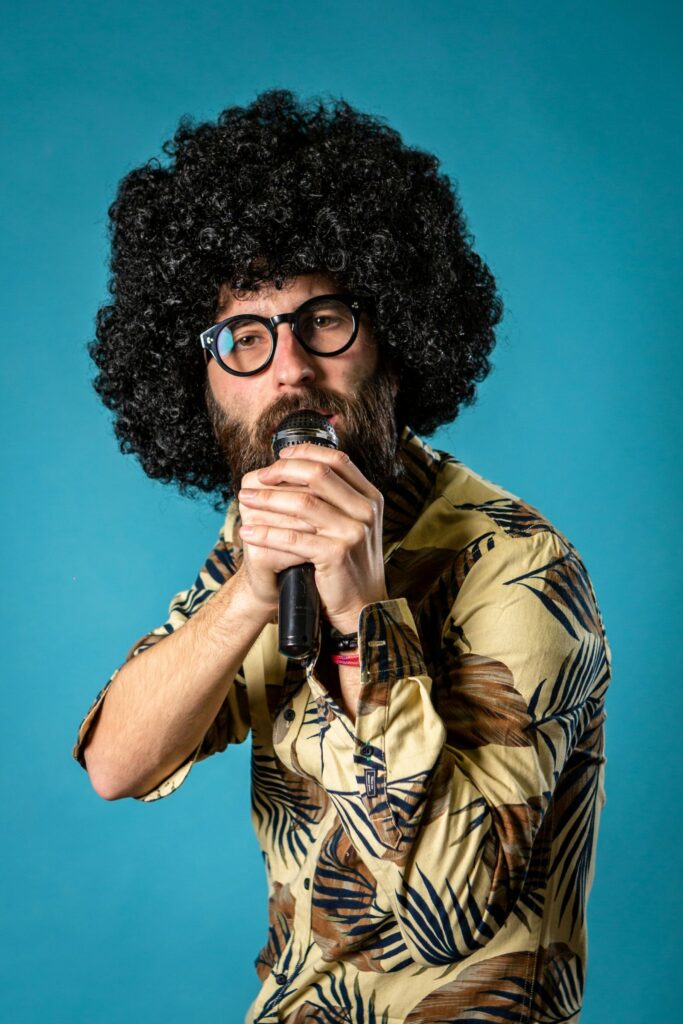 Stand-up comedian with an afro wig, beard and mustache, and glasses holds a mic up to his mouth